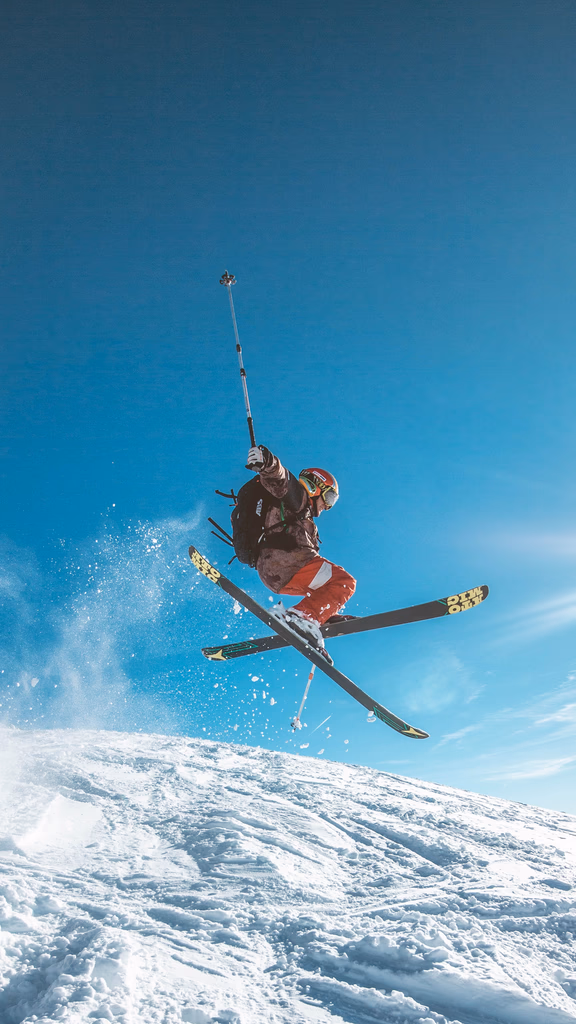 Skier in red pants and helmet jumping mid-air on a snowy slope under a clear blue sky.
