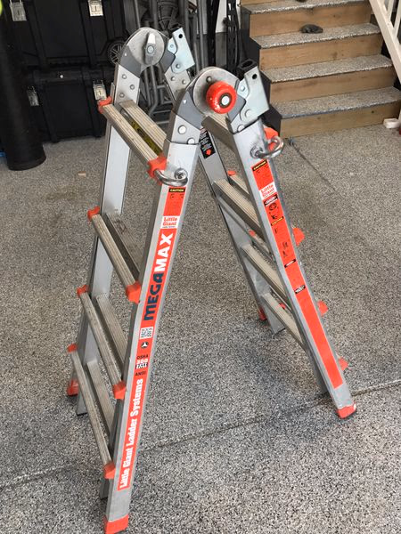 Silver folding multi-position ladder with red safety labels and rubber feet, standing open on a speckled gray floor.