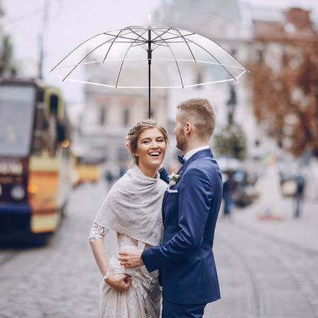 Smiling bride and groom standing close under a transparent umbrella on a city street.