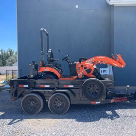 Orange compact tractor loaded on a black dual-axle trailer parked on gravel near a grey building.