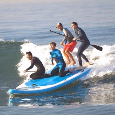 Four people surfing together on a large blue paddleboard riding a wave.