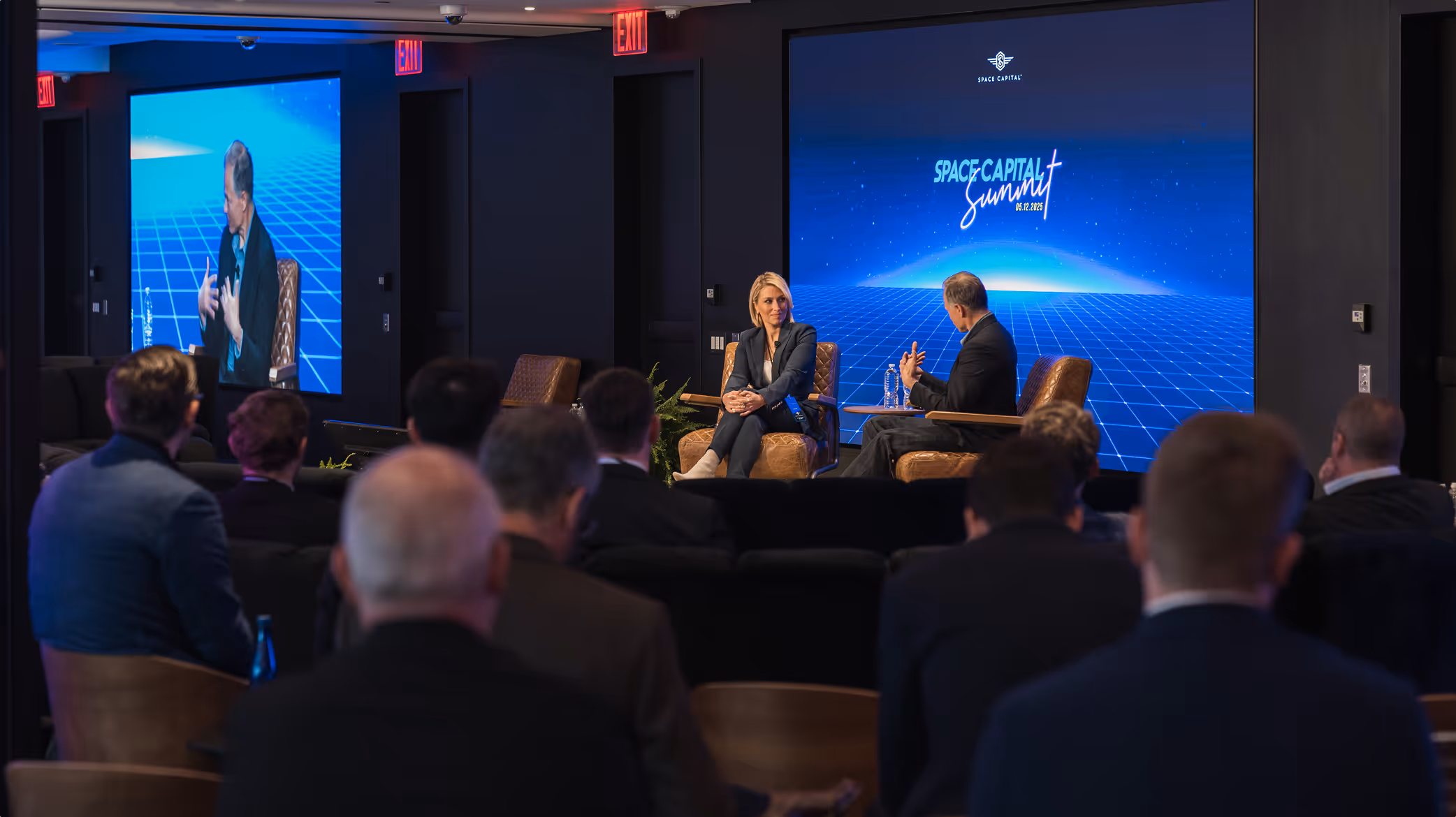 Panel discussion at the Space Capital Summit, showing two speakers on stage in front of a blue digital backdrop with grid lines and the event logo, while attendees watch from the audience.