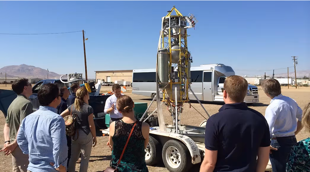 Group of people gathered outdoors around a small rocket prototype mounted on a trailer, listening to a presenter explaining its design at a desert testing facility.