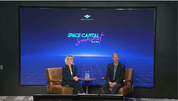 Two speakers seated on stage during the Space Capital Summit, with a large digital screen behind them displaying the event logo and a blue grid background inspired by futuristic design.