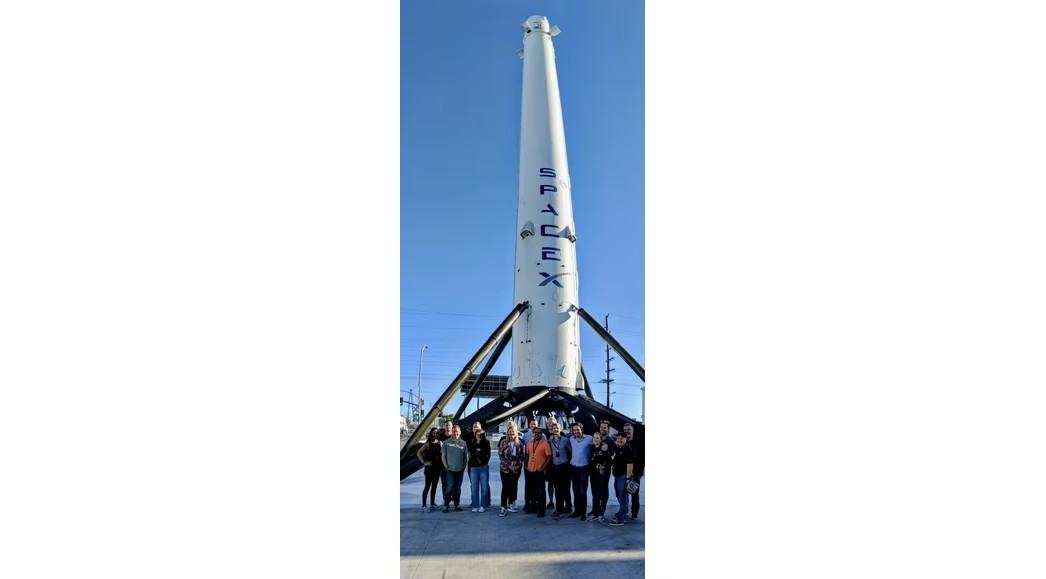 Group of people gathered outdoors around a small rocket prototype mounted on a trailer, listening to a presenter explaining its design at a desert testing facility.