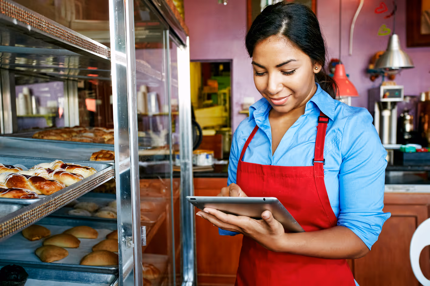 Bakery worker in red apron using tablet near display of fresh pastries