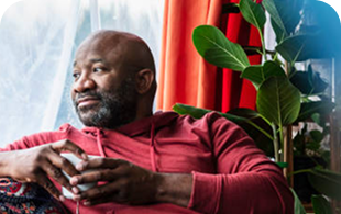 Man in red jacket leaning against window with potted plant beside him.