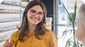 Woman wearing yellow shirt and glasses smiles in optical store with eyeglass display