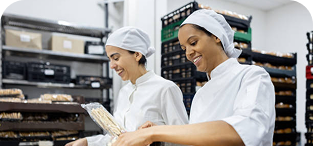 Two food industry workers in white uniforms and head coverings reviewing clipboard in bakery kitchen