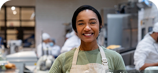Smiling woman wearing green apron and chef's hat in busy kitchen
