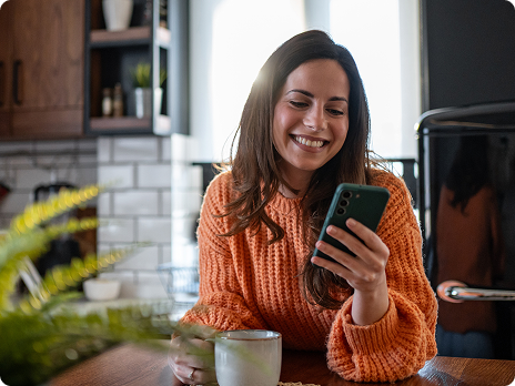 Smiling woman in orange sweater holding smartphone in modern kitchen.