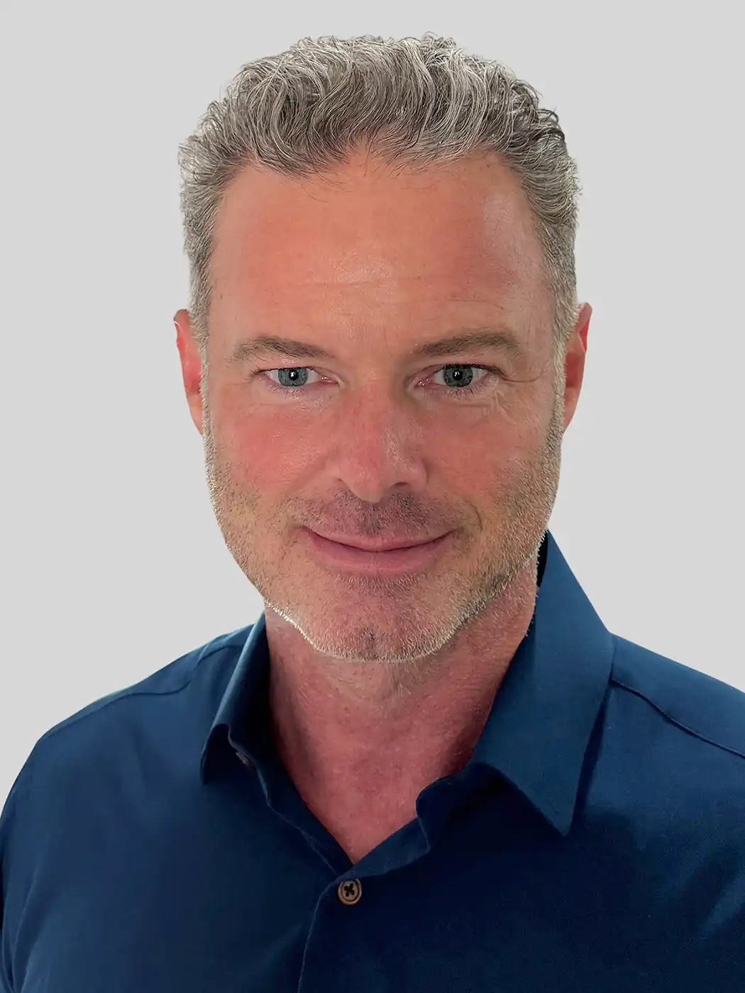 A headshot of a man smiling with silver hair wearing a dark blue button-up shirt