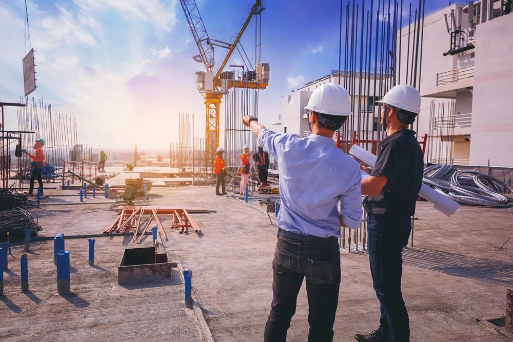 Two construction workers wearing white helmets discussing plans on a building site with cranes and other workers in the background during sunset.