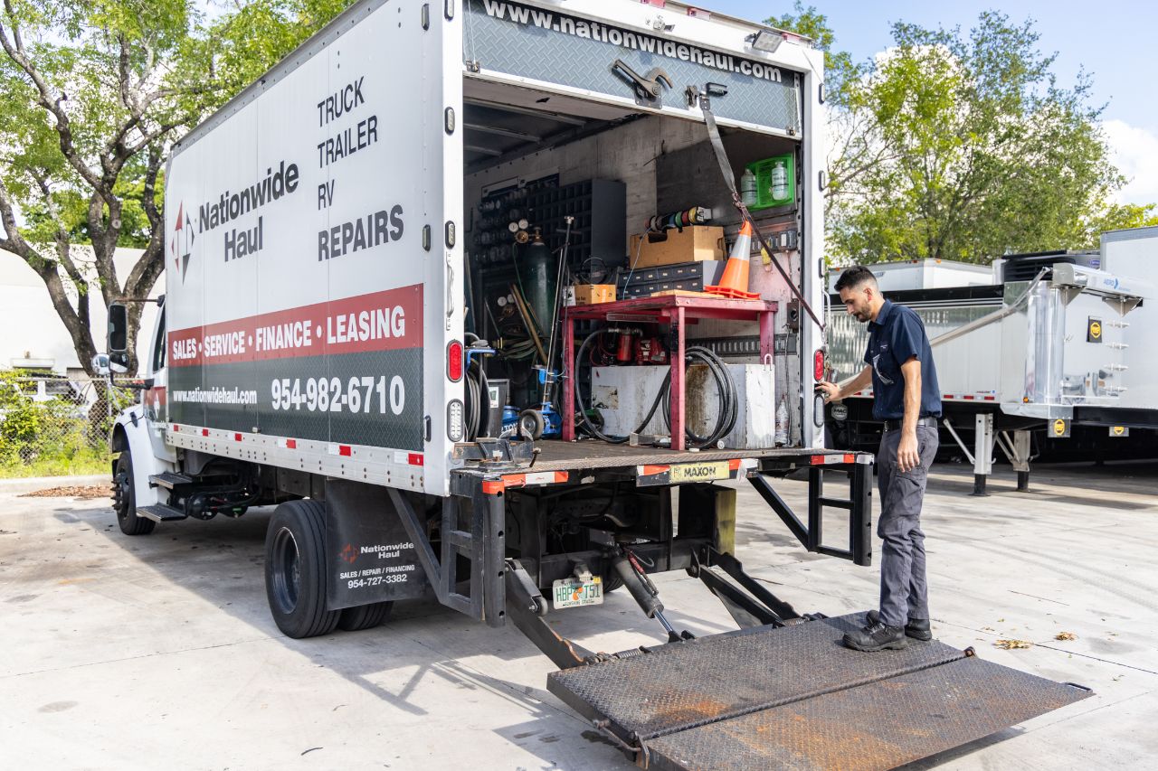 Mobile truck repair service vehicle with open rear compartment and liftgate as technician inspects equipment in outdoor lot beside trailers