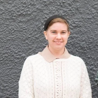 A lady in a white blouse with her hair pulled back stands smiling in front of a grey wall.