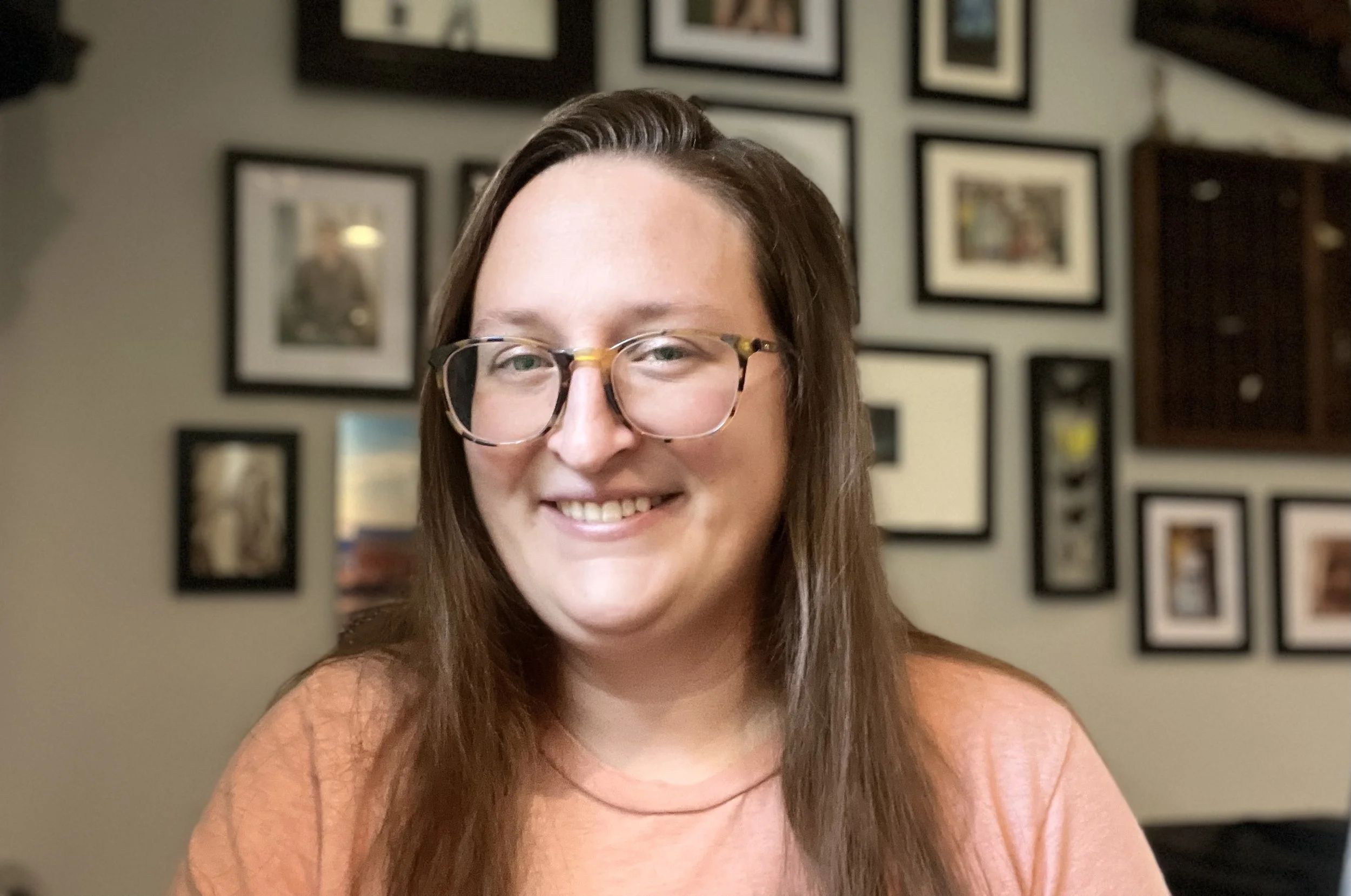 A lady in an orange t-shirt with long dark hair and glasses smiles in front of a wall of framed art.
