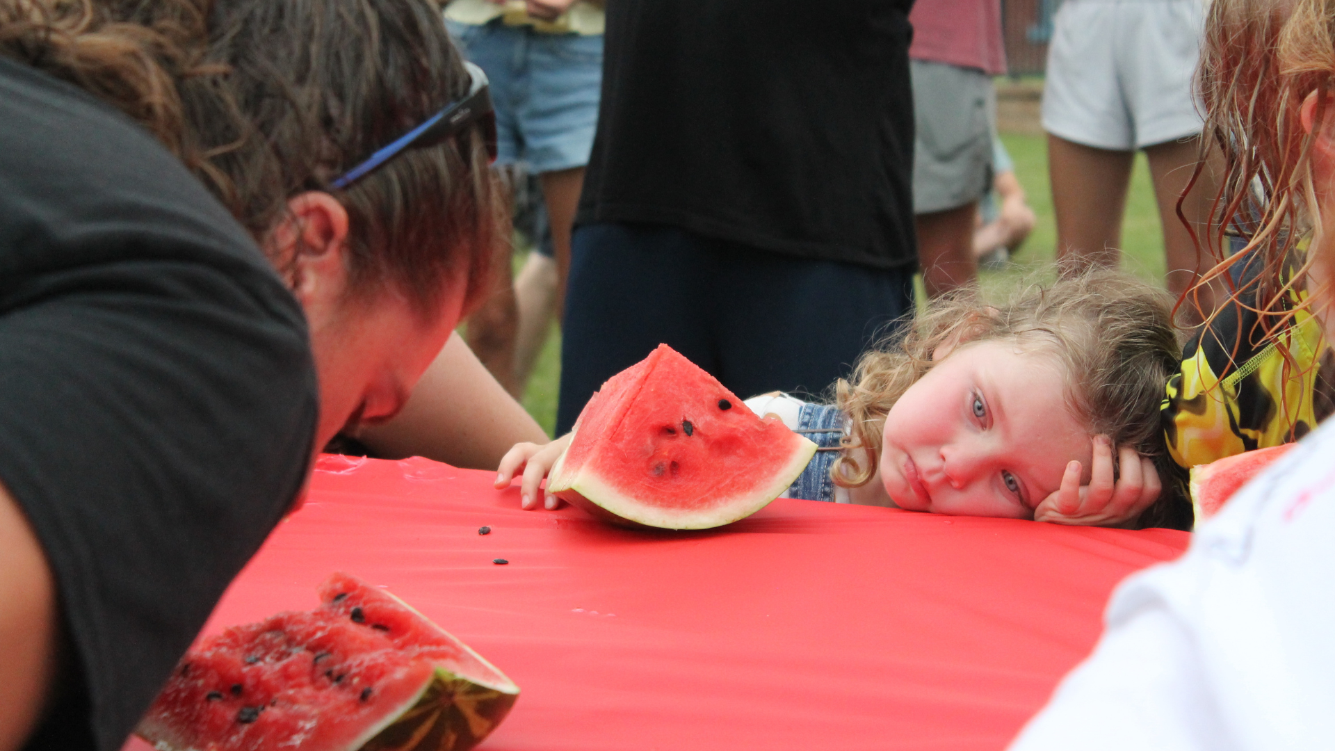 A young girl resting her head on a red table next to a slice of watermelon, with another person leaning close to the table and more people in the background.