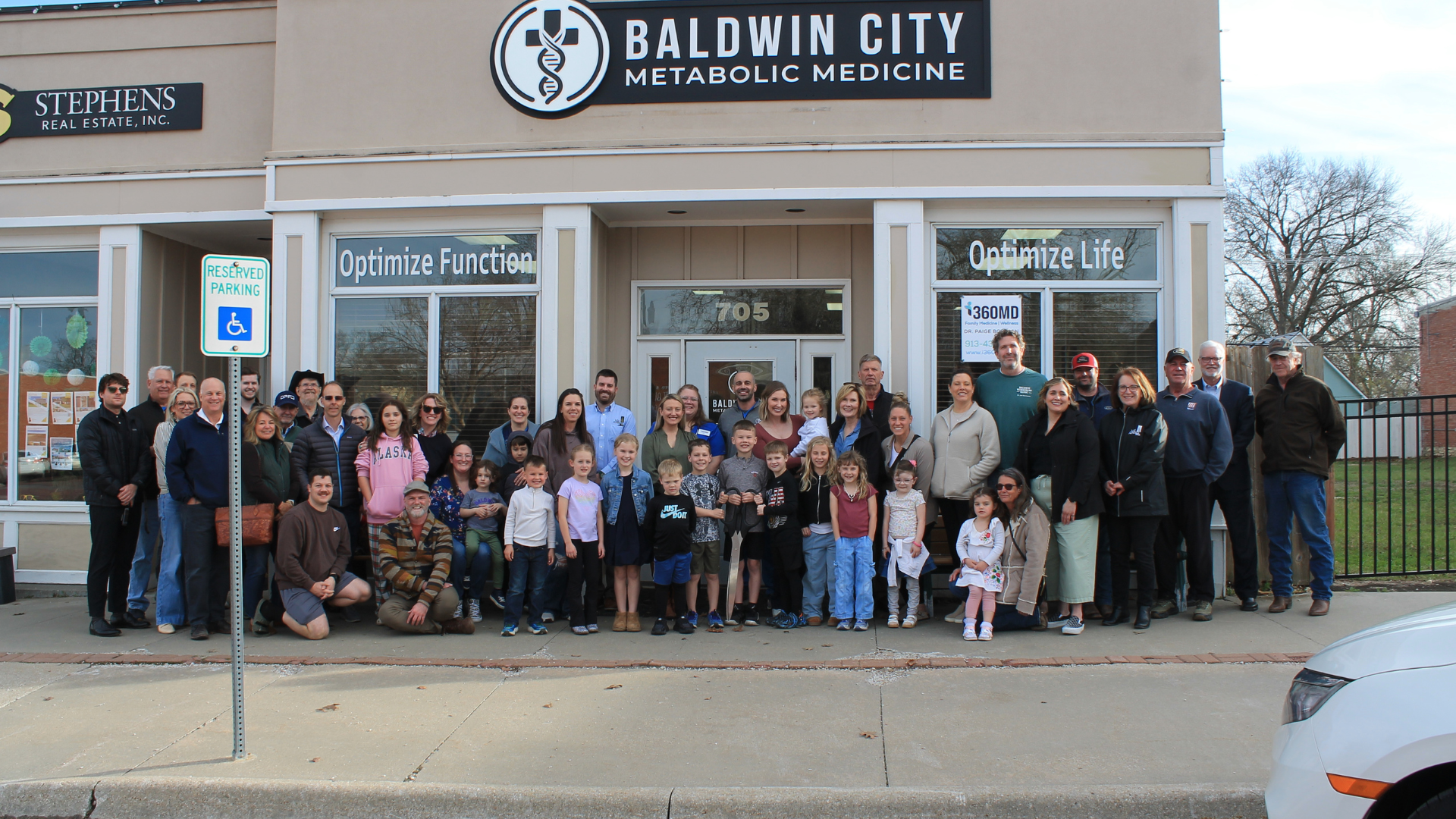 A large group of diverse people, including adults and children, pose for a photo outside Baldwin City Metabolic Medicine clinic.