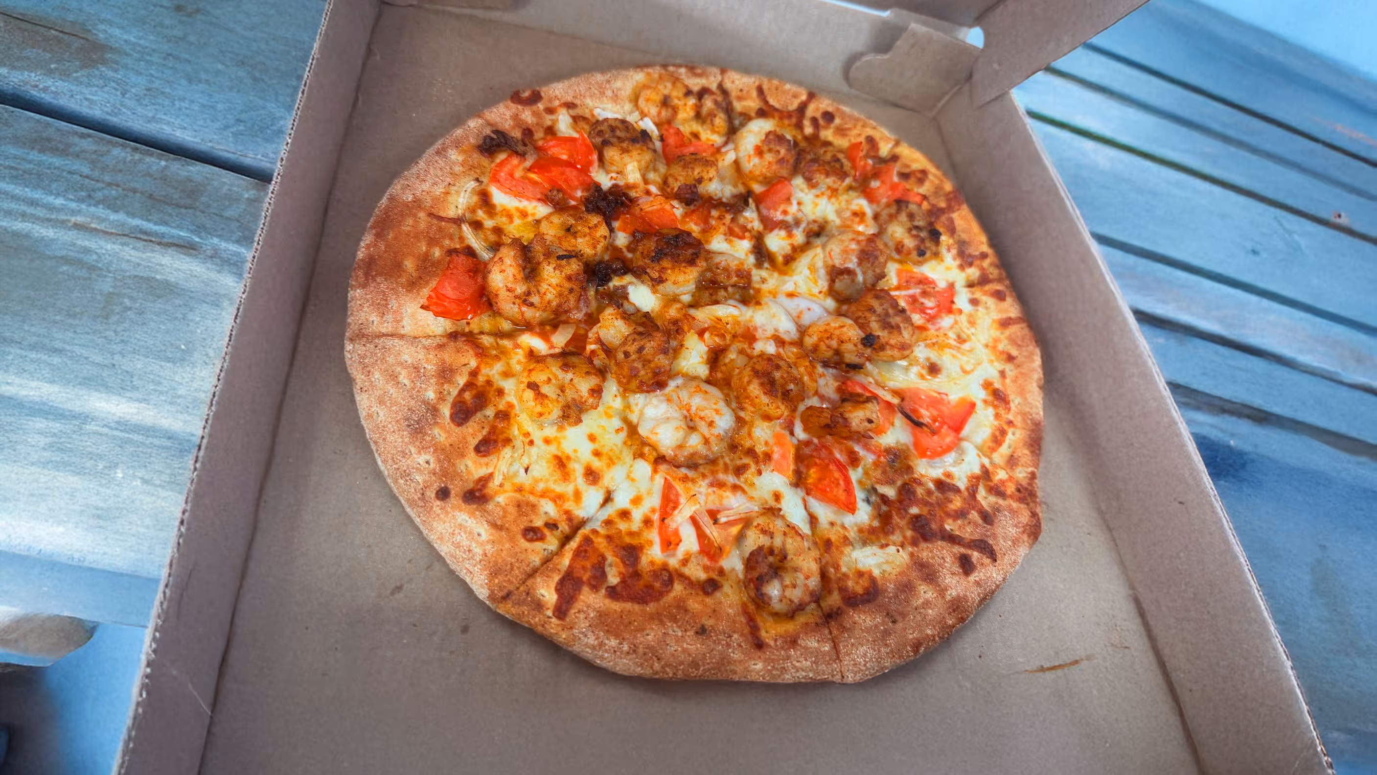 Pizza in an open box on a wooden table, topped with shrimp, melted cheese, and tomato pieces.