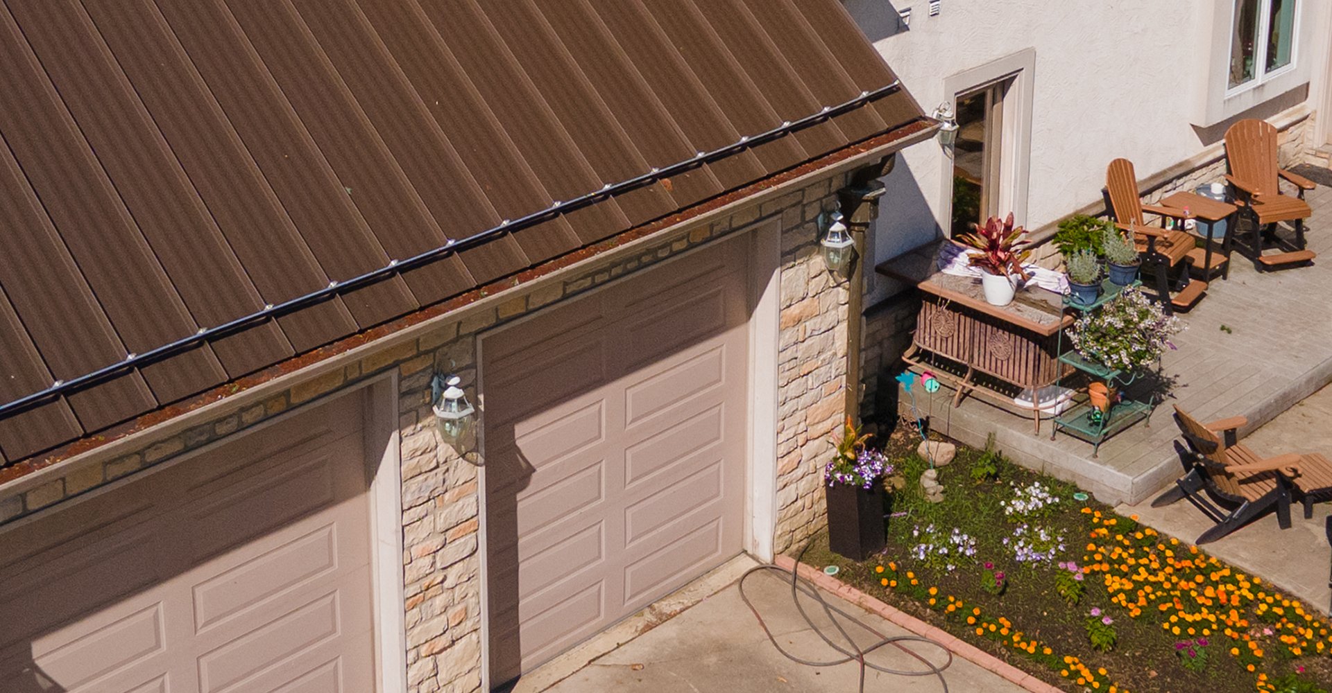 Aerial view of a metal roof edge with gutter and downspout, next to a two-car garage with stone accents and a paved patio with flowers and outdoor furniture
