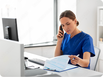 Female healthcare worker in blue scrubs on phone, reviewing medical documents at desk with computer.