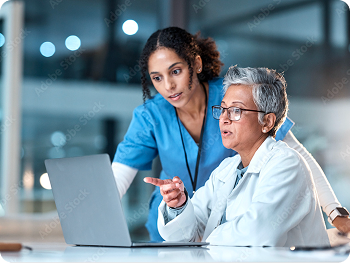 Older female doctor in white coat and younger nurse in blue scrubs discussing info on a laptop.