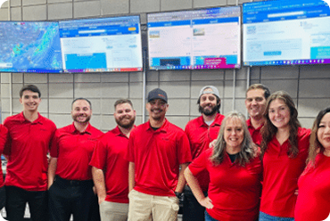 Group of nine people in red shirts standing in front of large monitors on a wall.