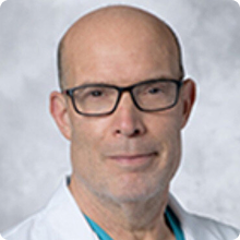 Bald man with glasses wearing a white medical coat against a neutral gray background.