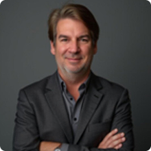 Smiling man in a dark blazer with crossed arms against a gray background.