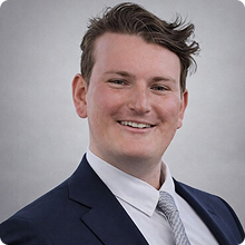 Smiling young man with short brown hair wearing a navy suit, white shirt, and gray tie on gray background.