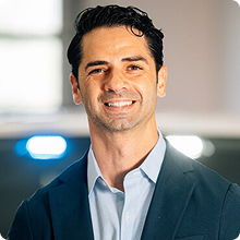 Smiling man with dark hair wearing a navy blazer and light blue shirt in an office setting.