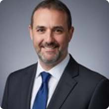 Smiling man with short hair and beard wearing a dark suit and blue tie against a gray background.