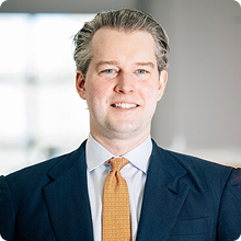 Smiling man in a suit with white shirt and orange tie in a bright indoor setting.