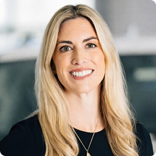 Smiling blonde woman with long hair wearing a black top and gold necklace in a blurred office background.