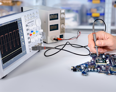 Hand testing electronics circuit board with oscilloscope showing waveform on screen.