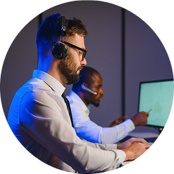 Two men wearing headsets working on computers in a dim office with blue lighting.