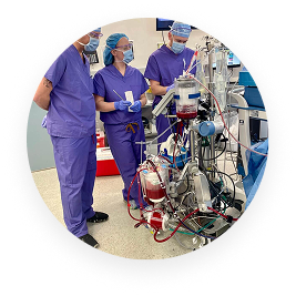 Three healthcare workers in blue scrubs operating a complex medical machine with tubes and blood bags.
