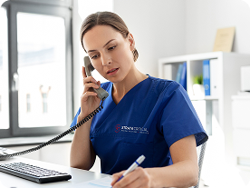 Female nurse in blue scrubs talking on phone while reviewing documents at a desk with computer.