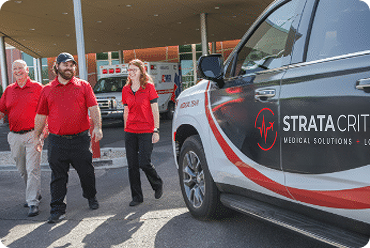 Three people in red shirts walking near a Trinity Medical Solutions vehicle outside a medical facility.