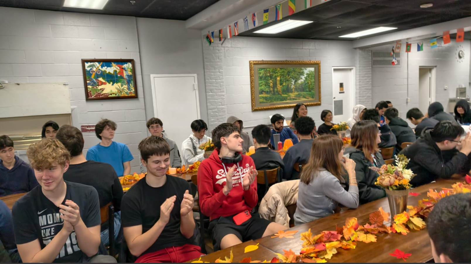 A diverse group of people seated at long tables decorated with autumn leaves and flower arrangements, some clapping and others engaged in conversation in a room with framed paintings and international flags.