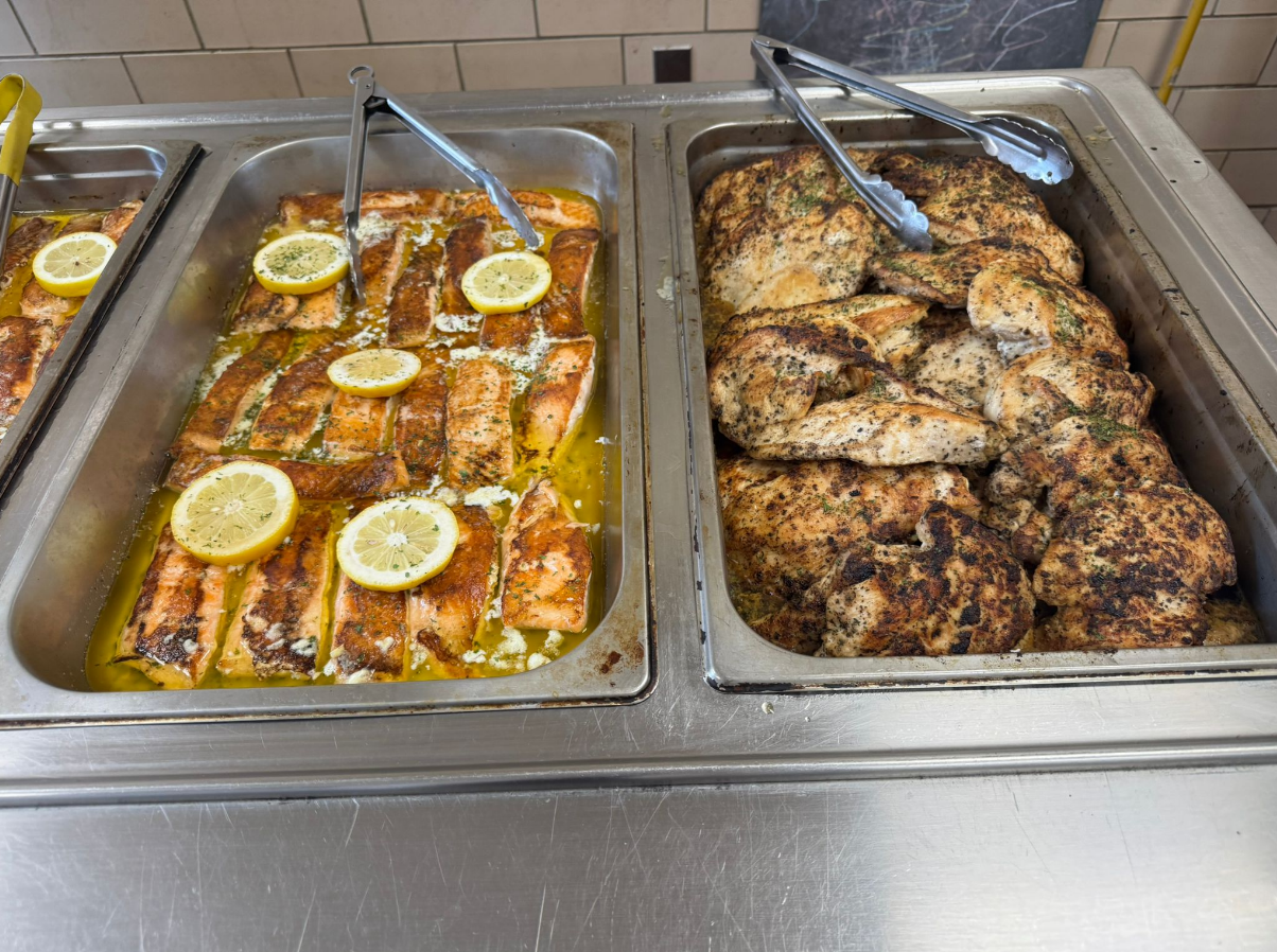 Buffet trays with cooked salmon fillets garnished with lemon slices and grilled seasoned chicken breast pieces with tongs.