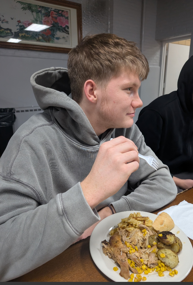 Young man in a gray hoodie sitting at a table holding a fork, with a plate of food including roasted potatoes, pulled meat, corn, and a dinner roll in front of him.