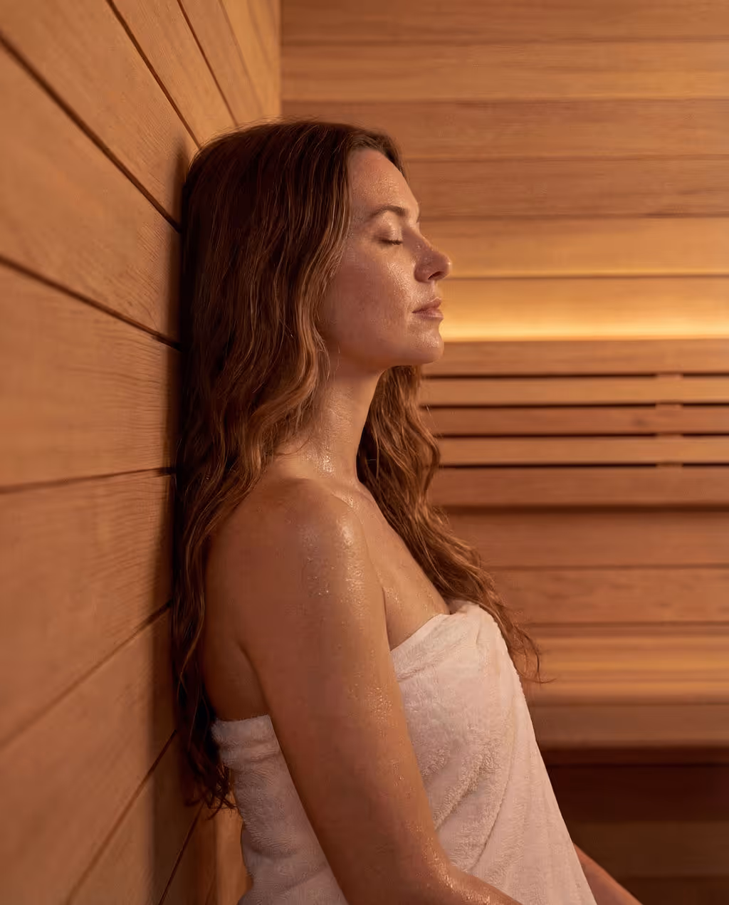 Woman wrapped in a towel relaxing with eyes closed against wooden wall inside a sauna.