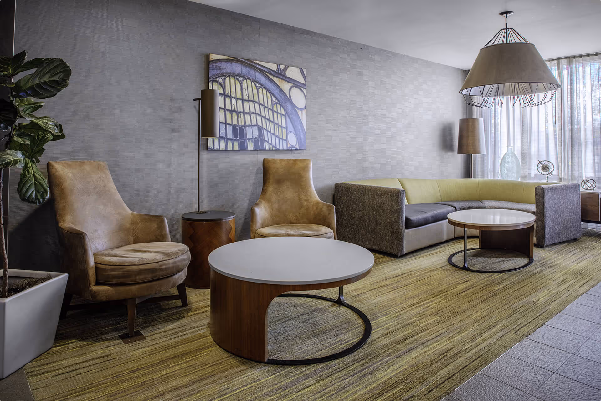Modern living room with two brown leather armchairs, a curved sectional sofa in green and gray, round coffee tables, floor lamps, and abstract wall art.