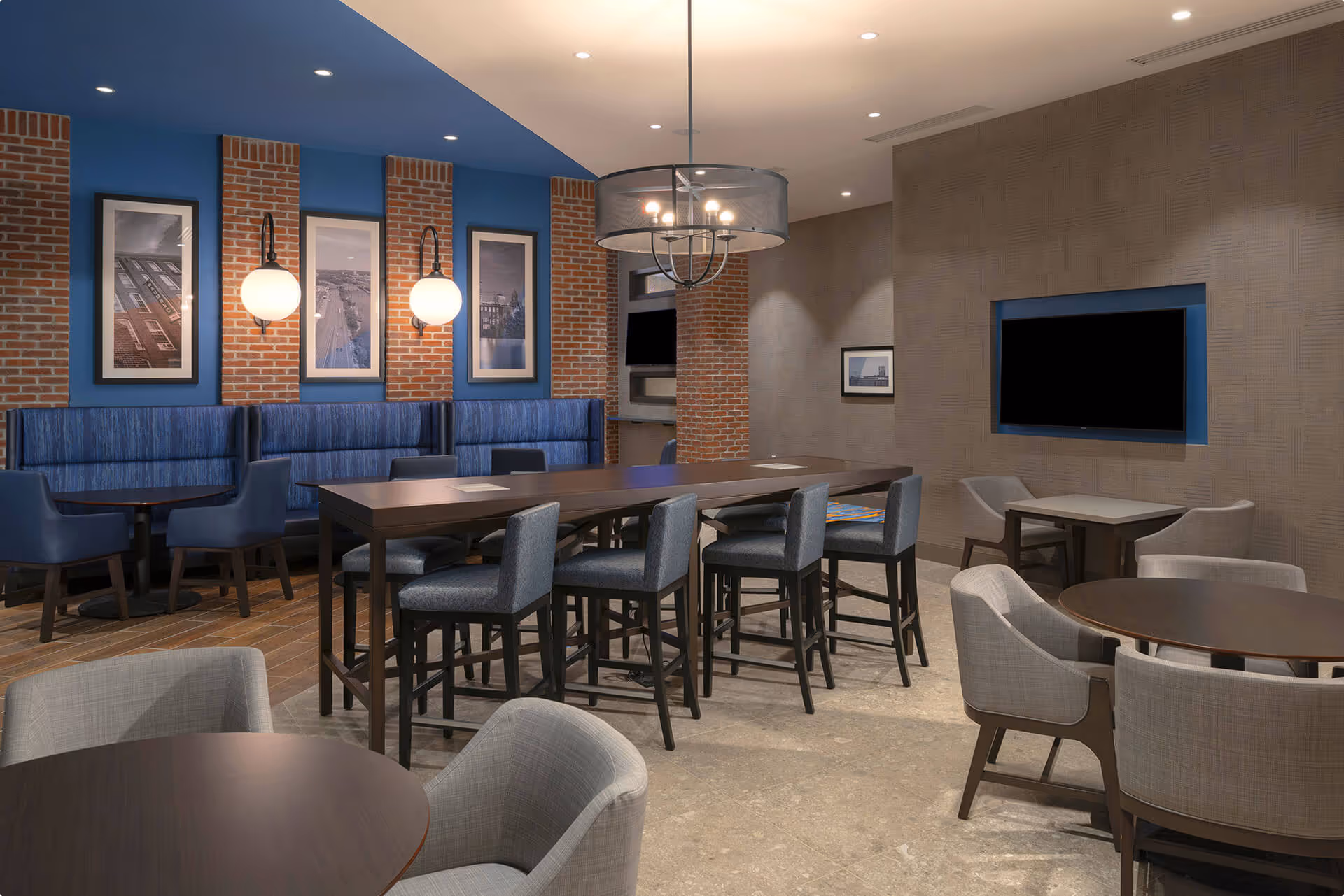 Modern hotel lounge with wooden tables, upholstered chairs, high stools around a long table, exposed brick columns, blue accent wall with framed black and white photos, and pendant lighting.