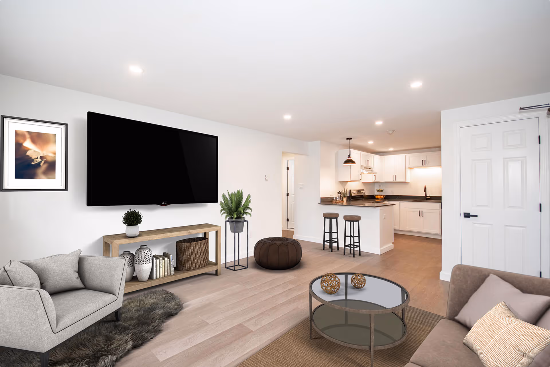 Modern open living room with a gray armchair, wall-mounted flat-screen TV, wooden console table with decor, round glass coffee table, and adjoining kitchen with white cabinets and bar stools.