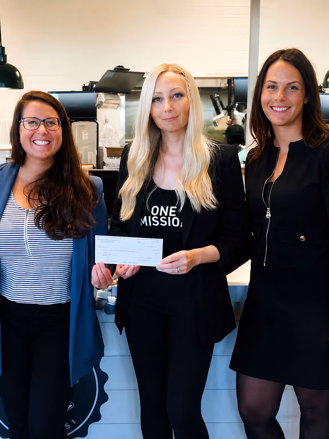 Three women standing indoors, with the middle woman holding a check and wearing a shirt that says 'ONE MISSION.'