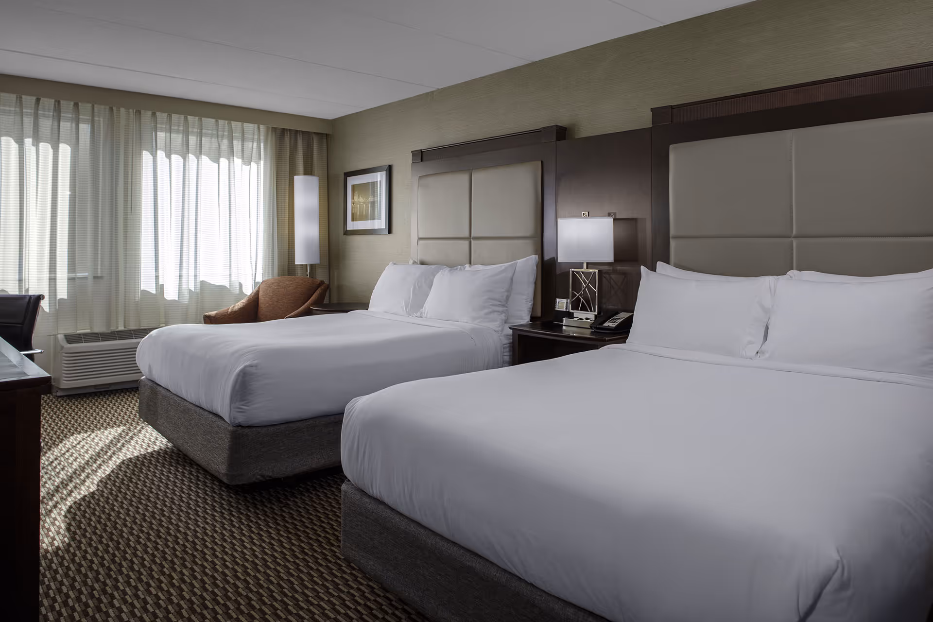 Hotel room with two queen beds, white linens, beige padded headboards, a nightstand with a lamp and phone, a brown chair near a window with sheer curtains.