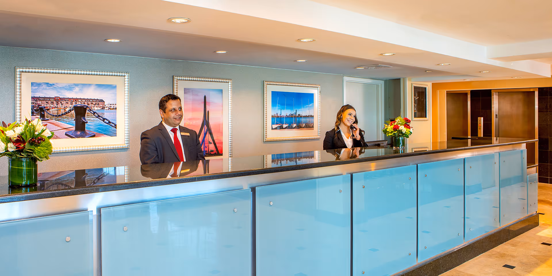 Two front desk staff in formal attire standing behind a modern hotel reception desk with framed pictures and flower arrangements.