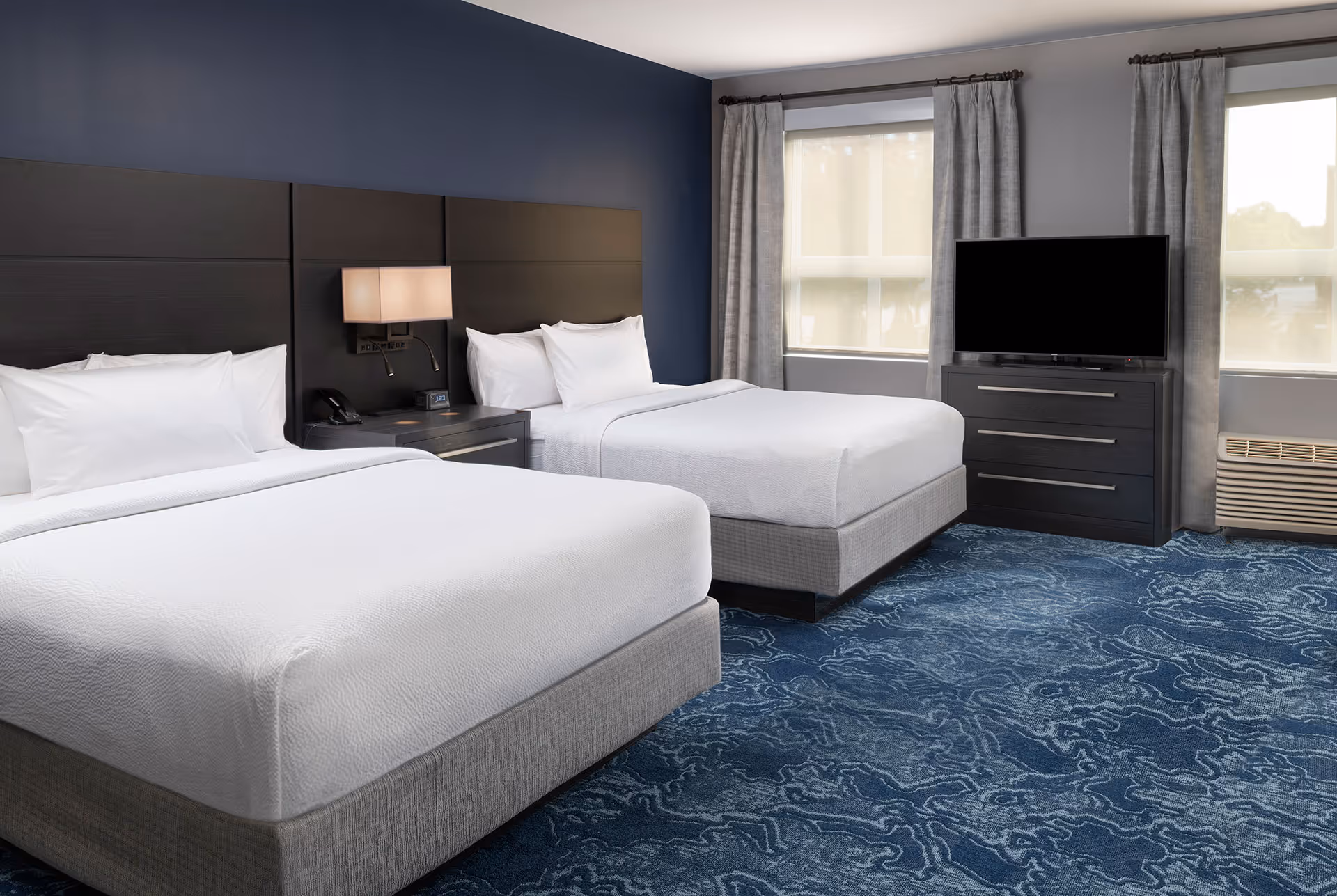 Hotel room with two queen beds, dark wood headboards, nightstand with lamp, TV on dresser, and blue patterned carpet.